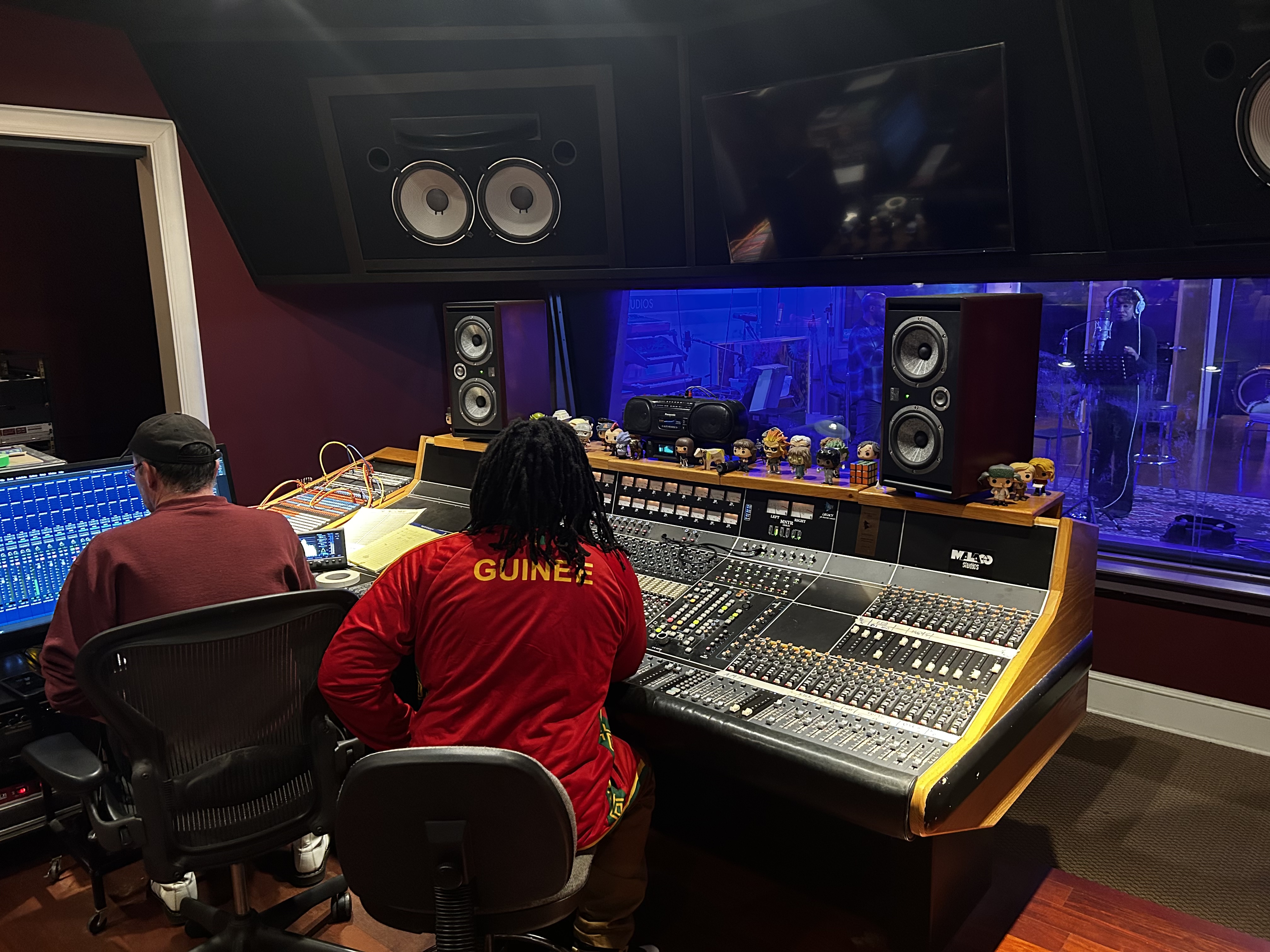 Baba Eddie in a red Guinée jacket seated at a large studio mixing console beside an engineer, a vocalist visible through the glass in the blue-lit live room