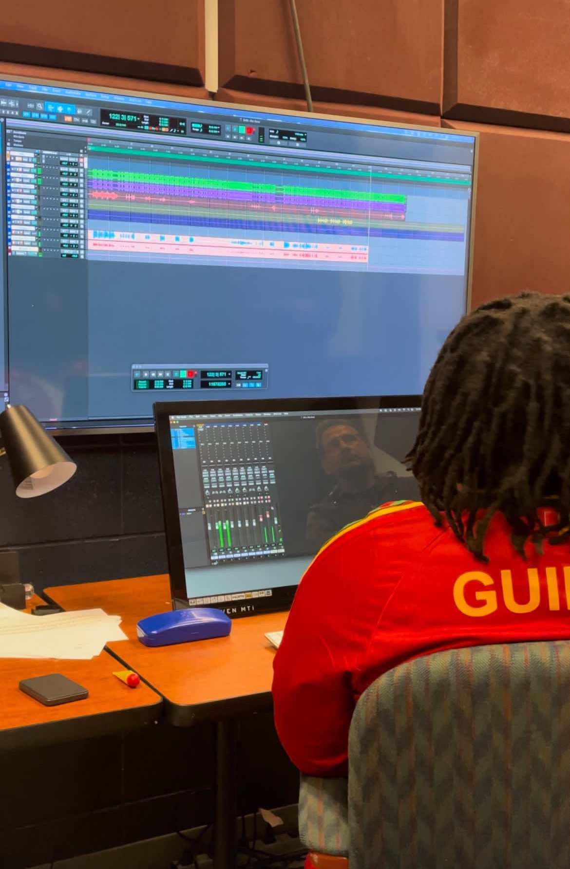 Baba Eddie in a red Guinée jersey at a studio desk viewing a digital audio workstation session with multi-track waveforms on a large screen