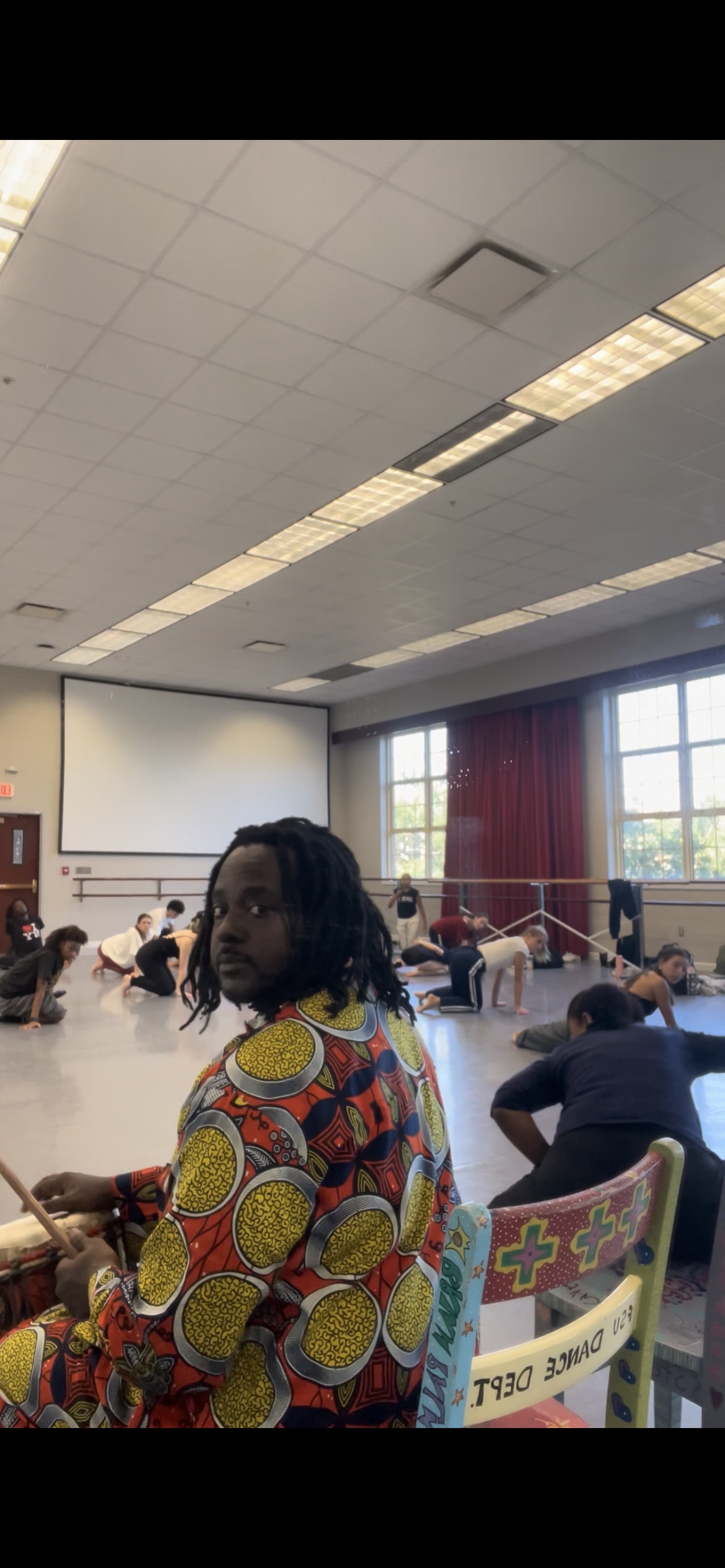 Baba Eddie in a vibrant orange and yellow West African print outfit drumming while dancers stretch and rehearse across a dance studio floor behind him