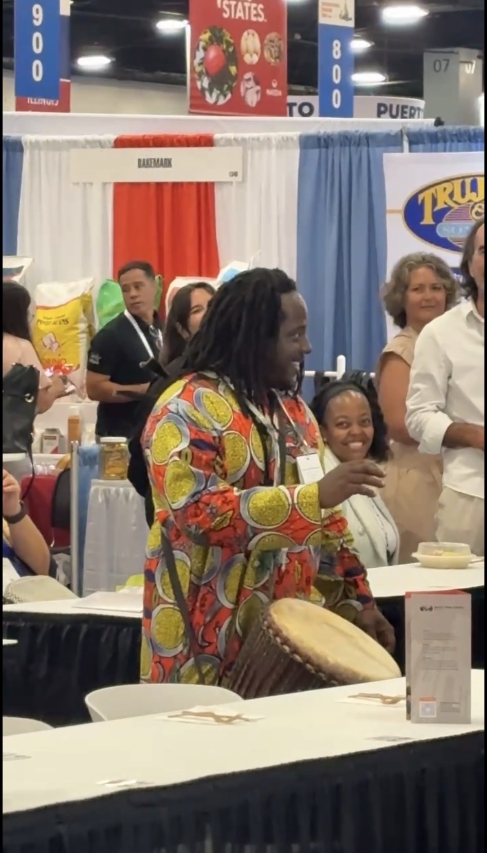 Baba Eddie playing a djembe at a lively expo booth as people gather around him smiling and engaging with the rhythm