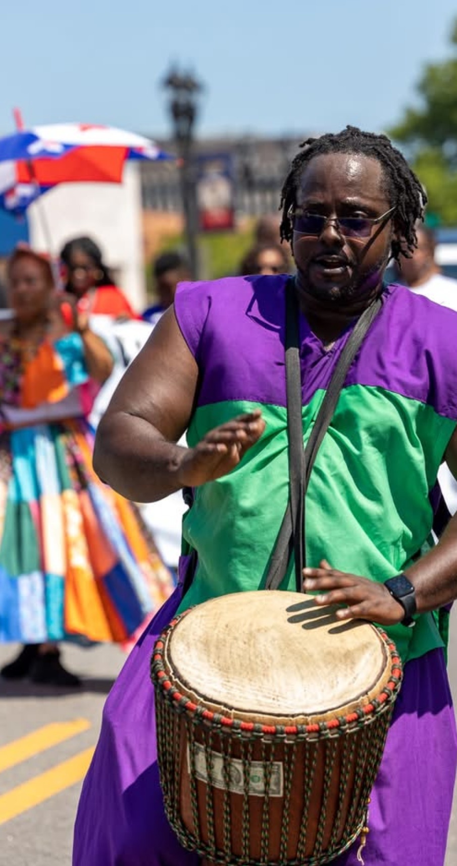 Baba Eddie playing a strapped djembe in a street parade, with dancers in patchwork skirts behind him