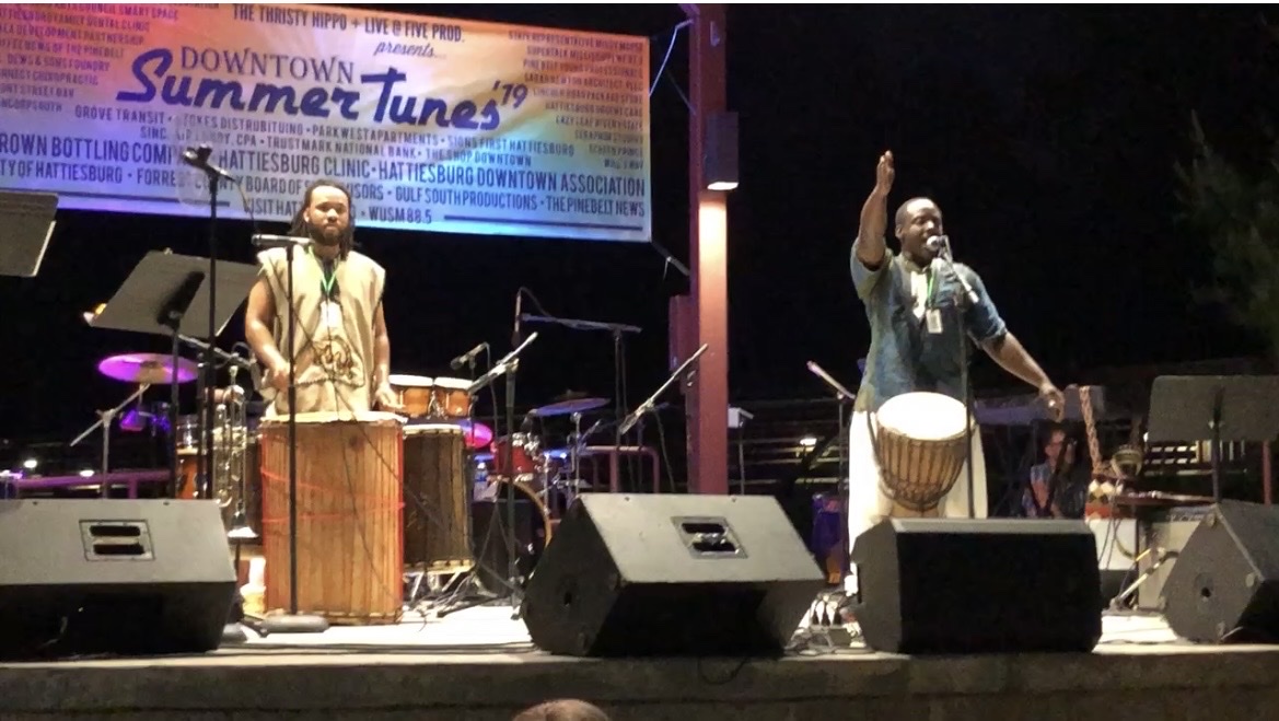 Baba Eddie and a fellow performer on an outdoor festival stage at night, one drumming and one singing with an arm raised, beneath a Downtown Summer Tunes banner