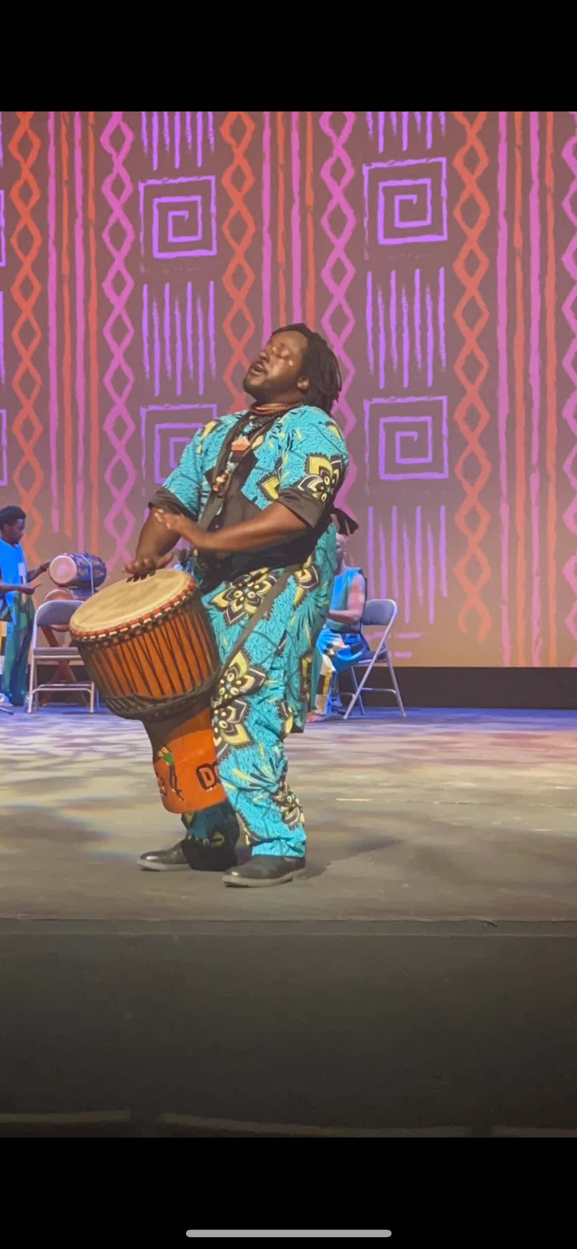 Baba Eddie arched back mid-strike on a strapped djembe on a theater stage against a patterned backdrop
